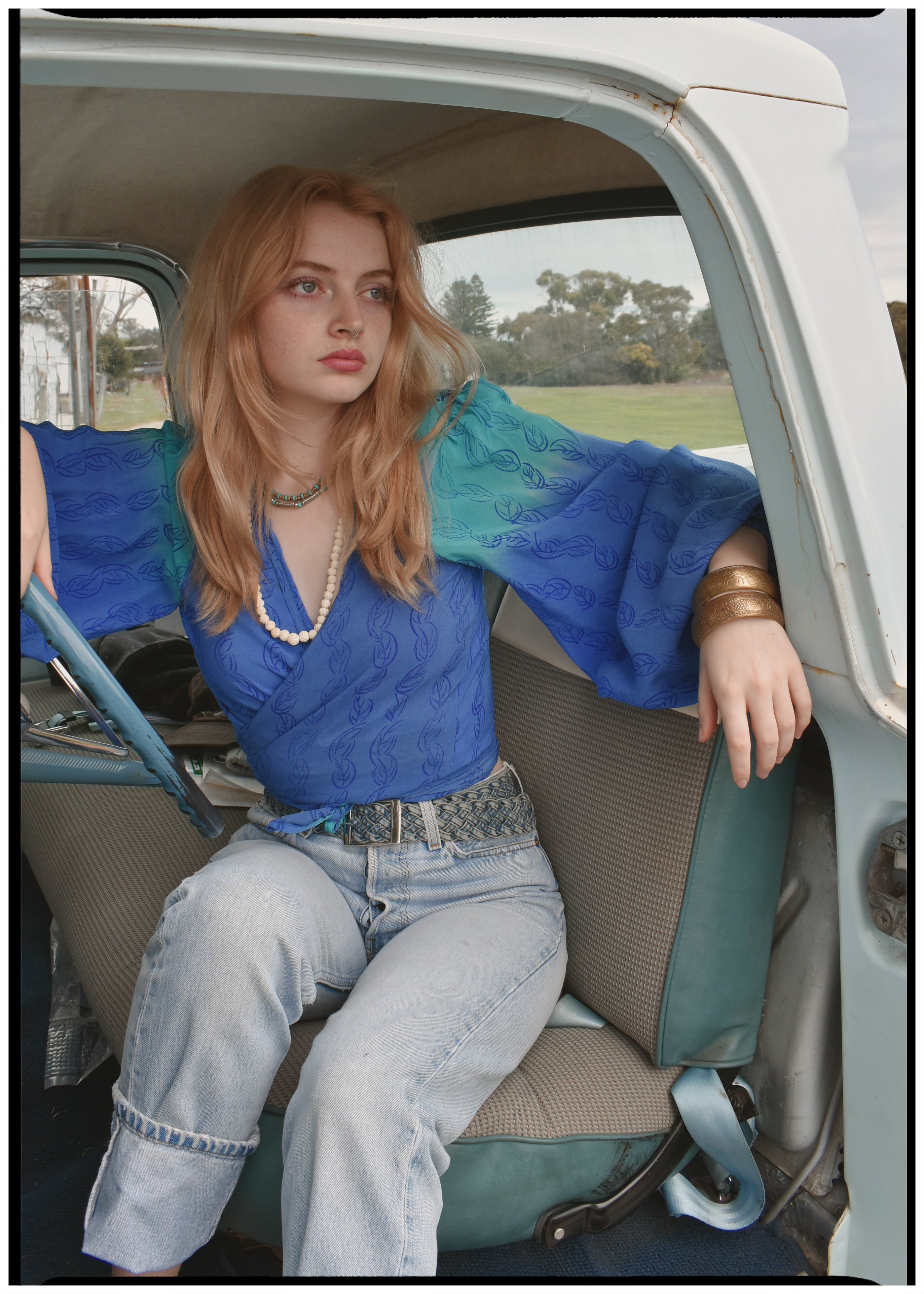 Woman wearing a bohemian style balloon sleeved blouse made from vintage Indian repurposed silk crepe saree sitting in the back of a vintage car with a scenic background