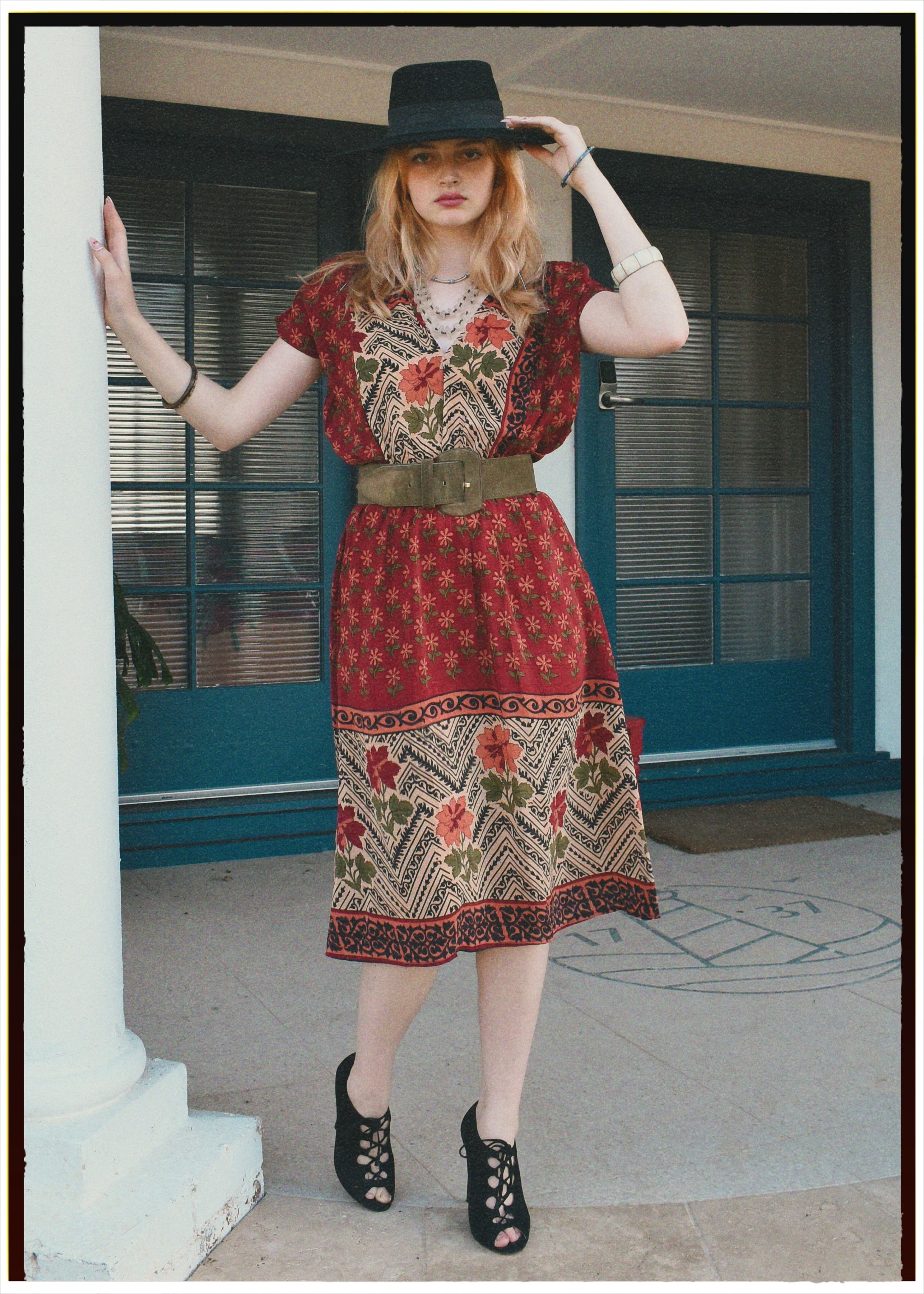 Woman wearing a patterned dress and hat, standing on a porch.