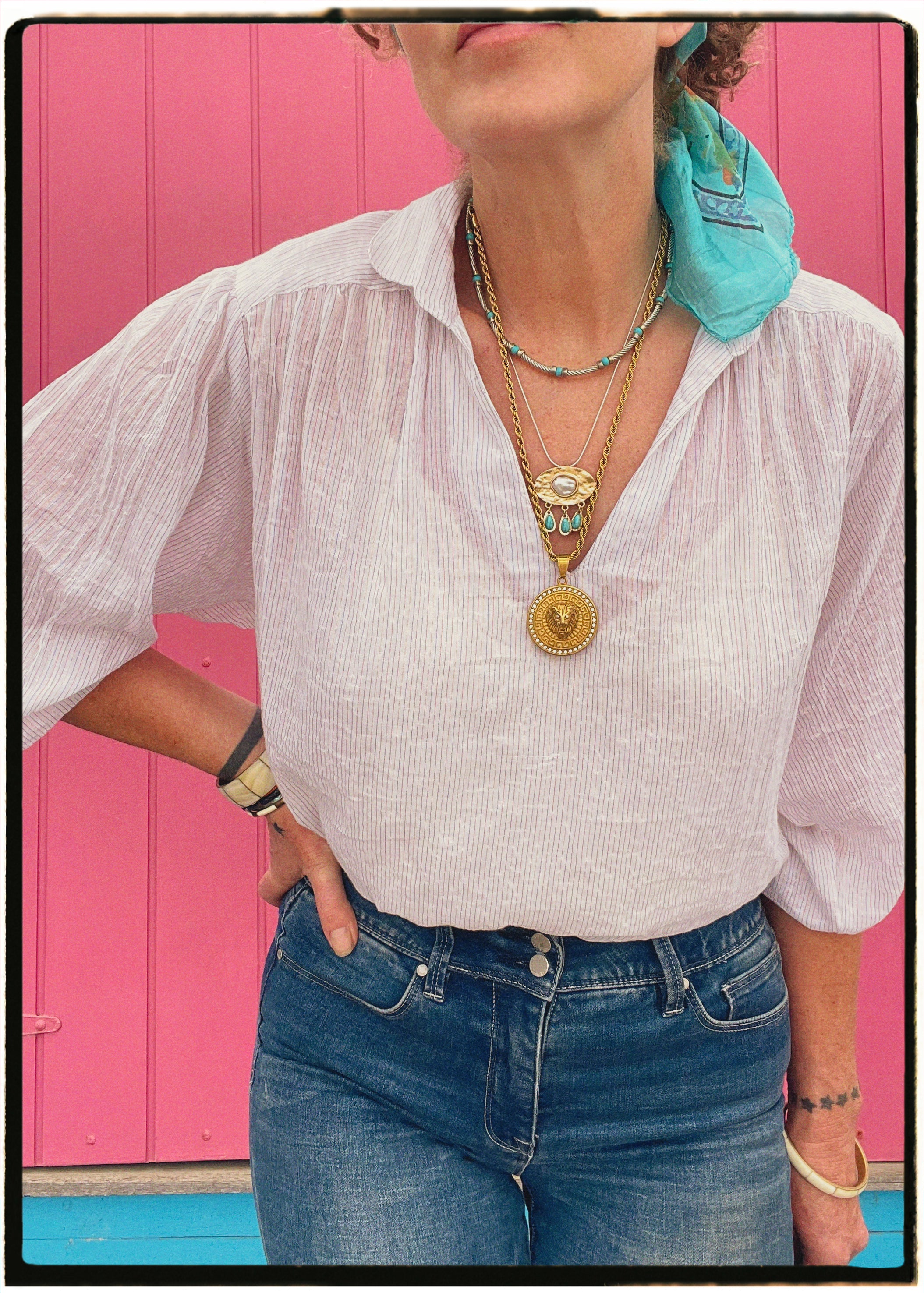 Person wearing a white shirt, blue jeans, and multiple necklaces with a pink background