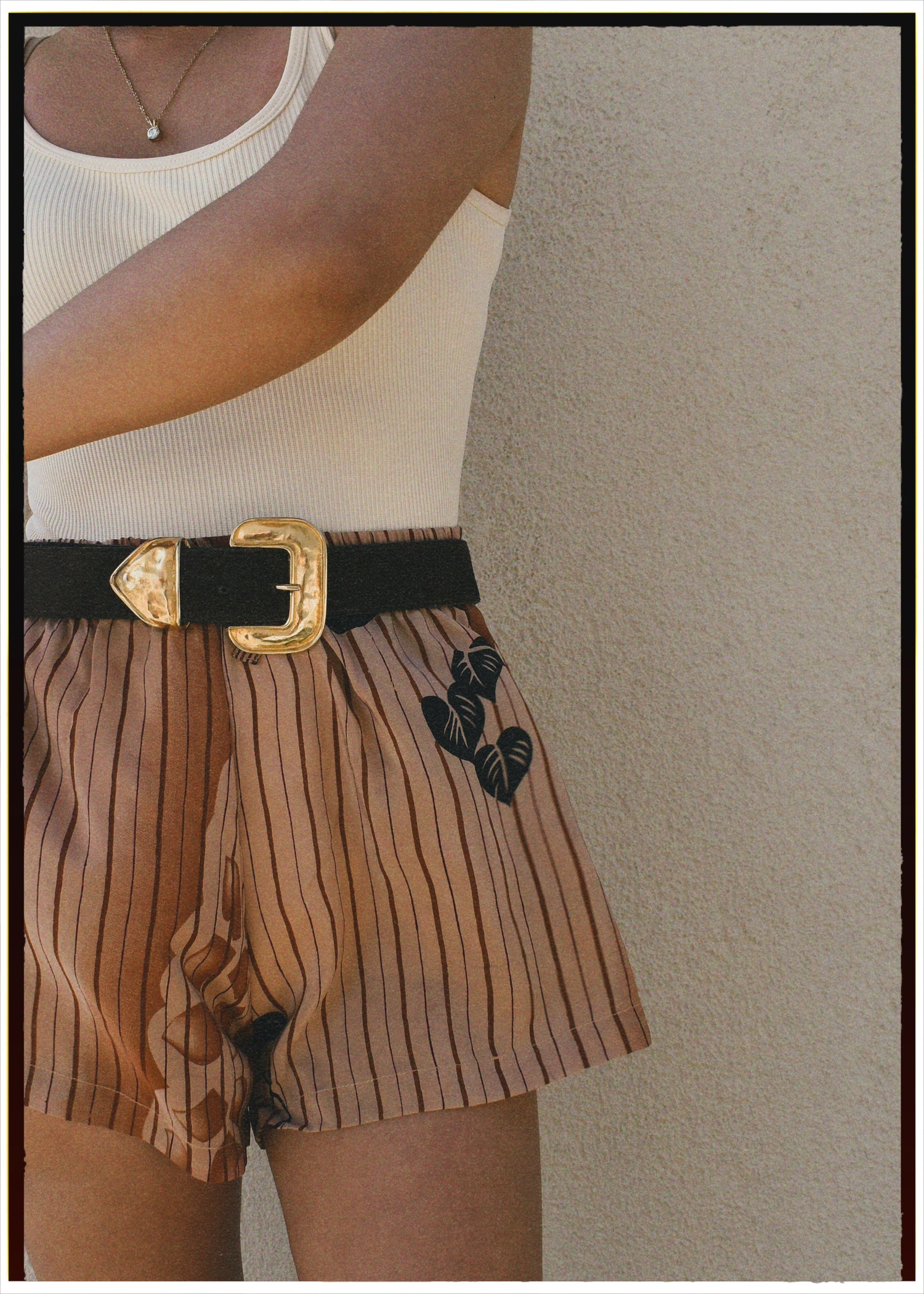 Person wearing a white sleeveless top and brown shorts with black belt and floral design on a neutral background