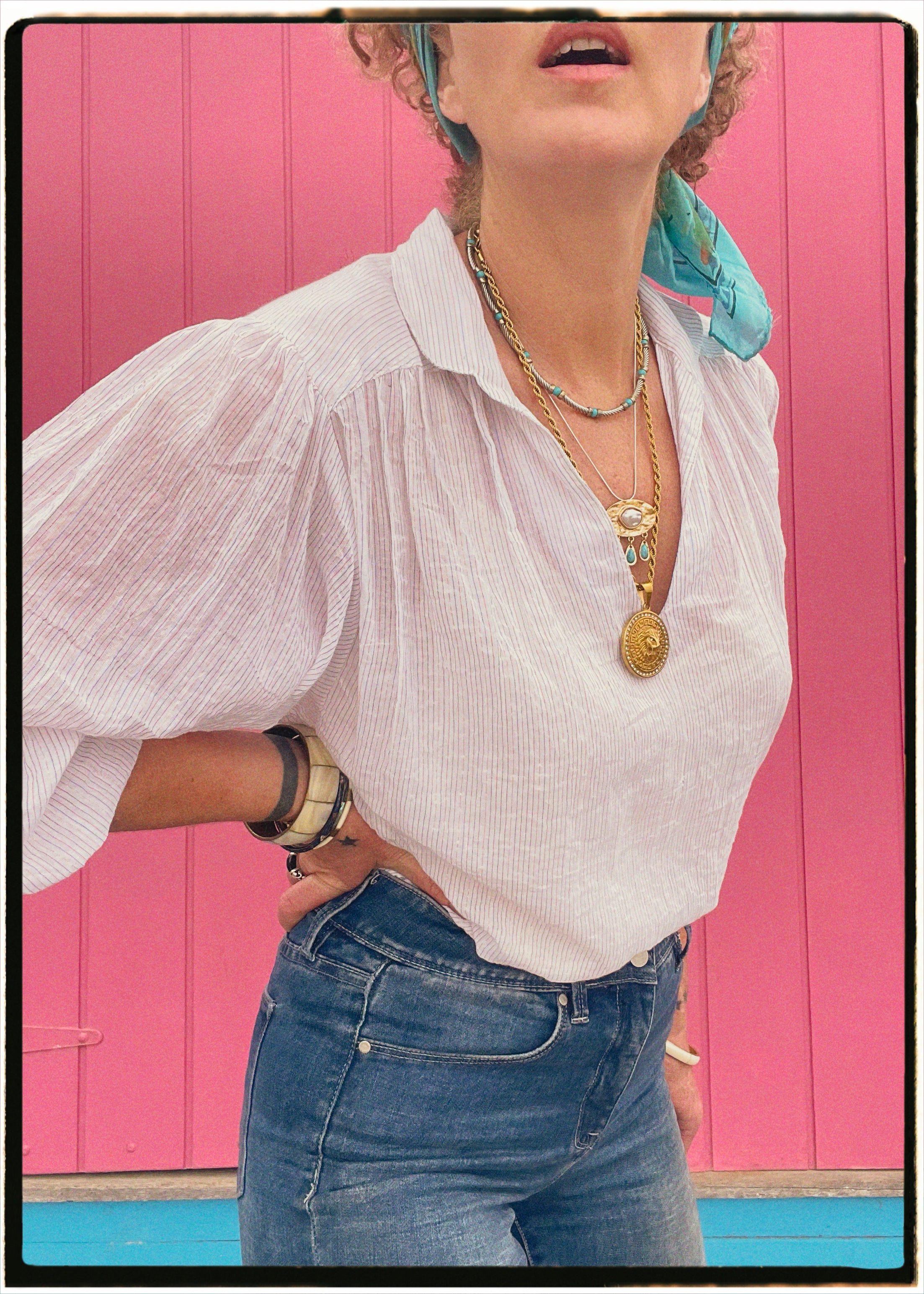 Person wearing a white blouse, blue jeans, and multiple necklaces against a pink background