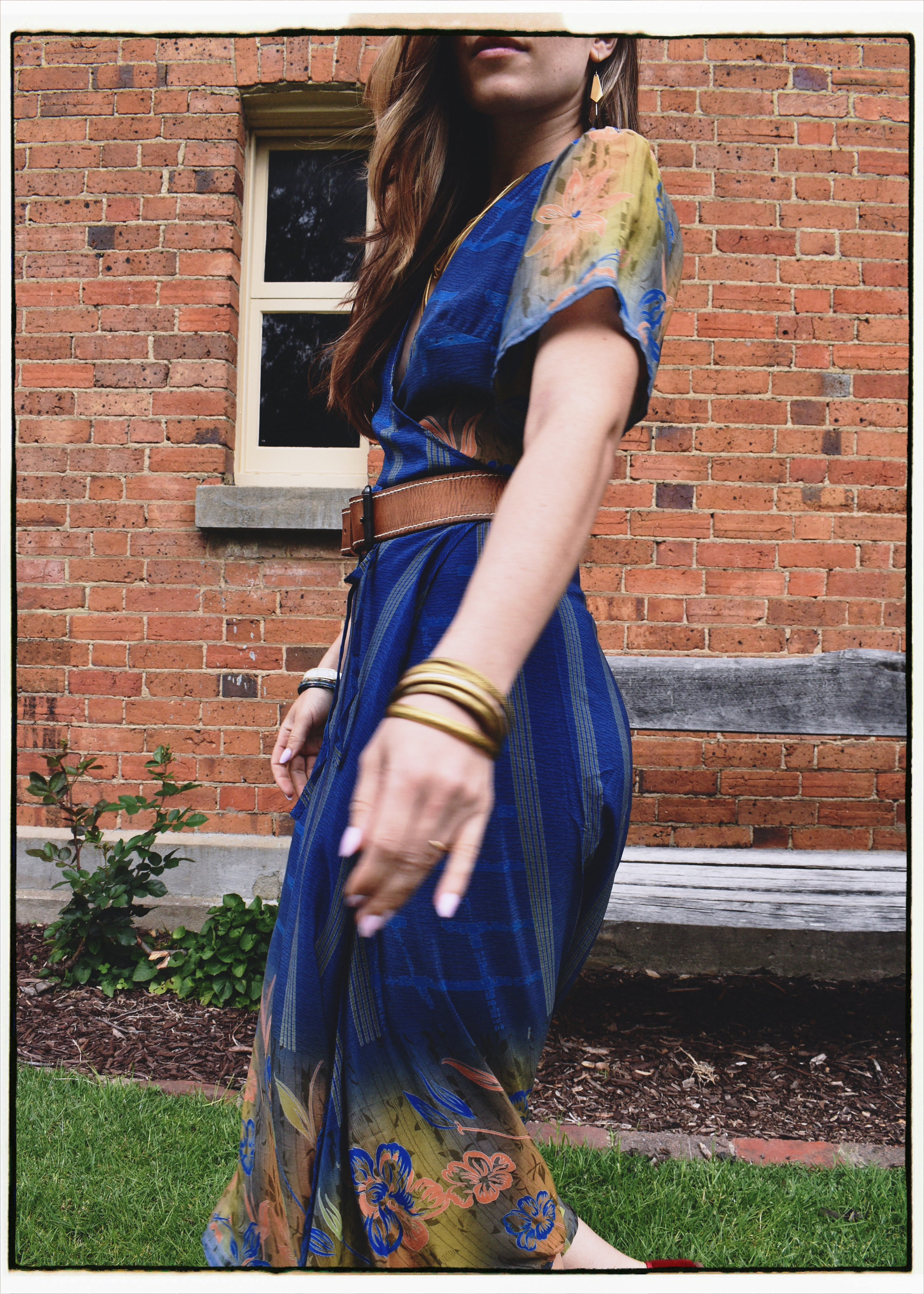 A woman wearing a blue and yellow floral maxi dress with a brown belt, standing outdoors in front of a red brick building.