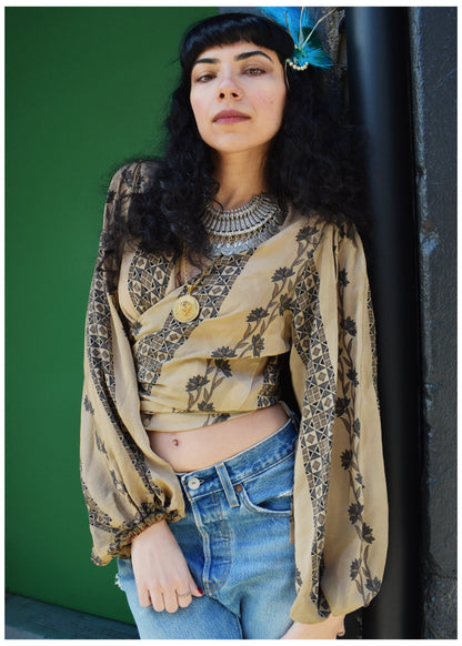 A woman with dark curly hair and a blue feather accessory wears a beige and black patterned bohemian long-sleeve crop top with sheer balloon sleeves, paired with blue jeans, posing against a green wall.