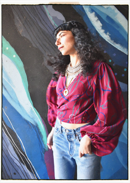 A woman with long curly black hair is wearing a vibrant magenta-red long-sleeve crop top with blue abstract line patterns and light-wash blue jeans, standing in front of a colorful abstract painted wall.