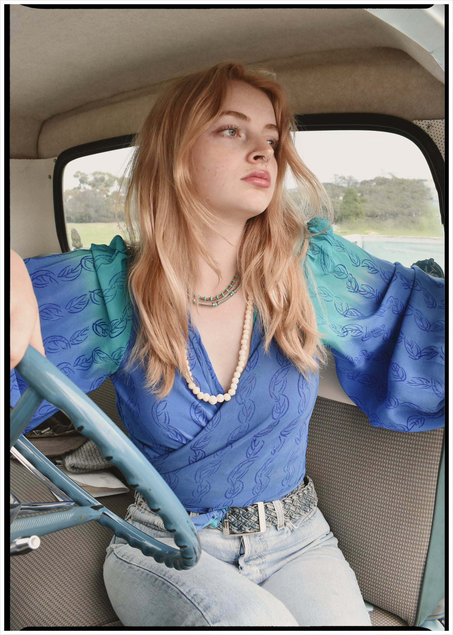 Woman wearing a bohemian style balloon sleeved blouse made from vintage Indian repurposed silk crepe saree sitting in the back of a vintage car with a scenic background