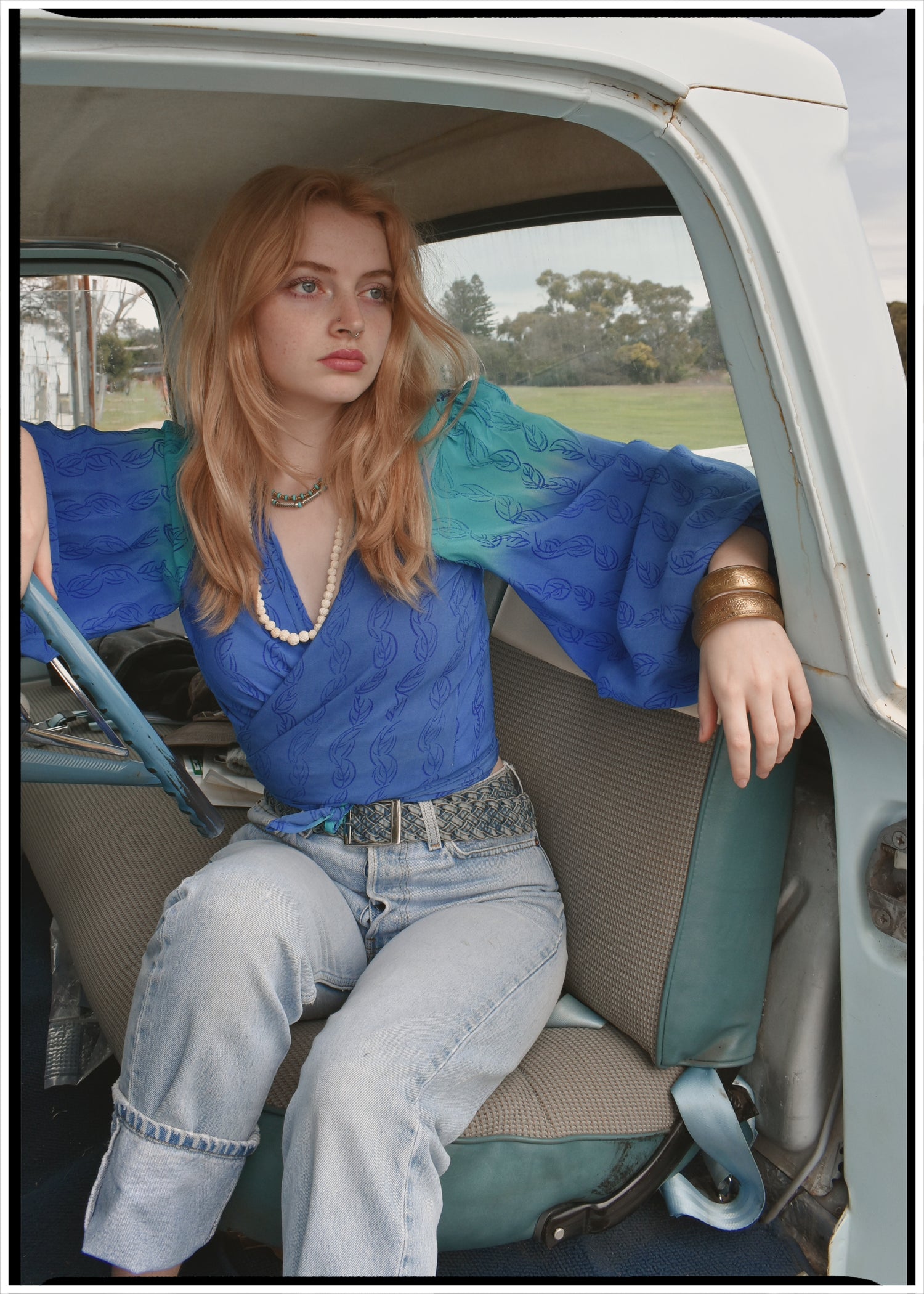 Woman wearing a bohemian style balloon sleeved blouse made from vintage Indian repurposed silk crepe saree sitting in the back of a vintage car with a scenic background