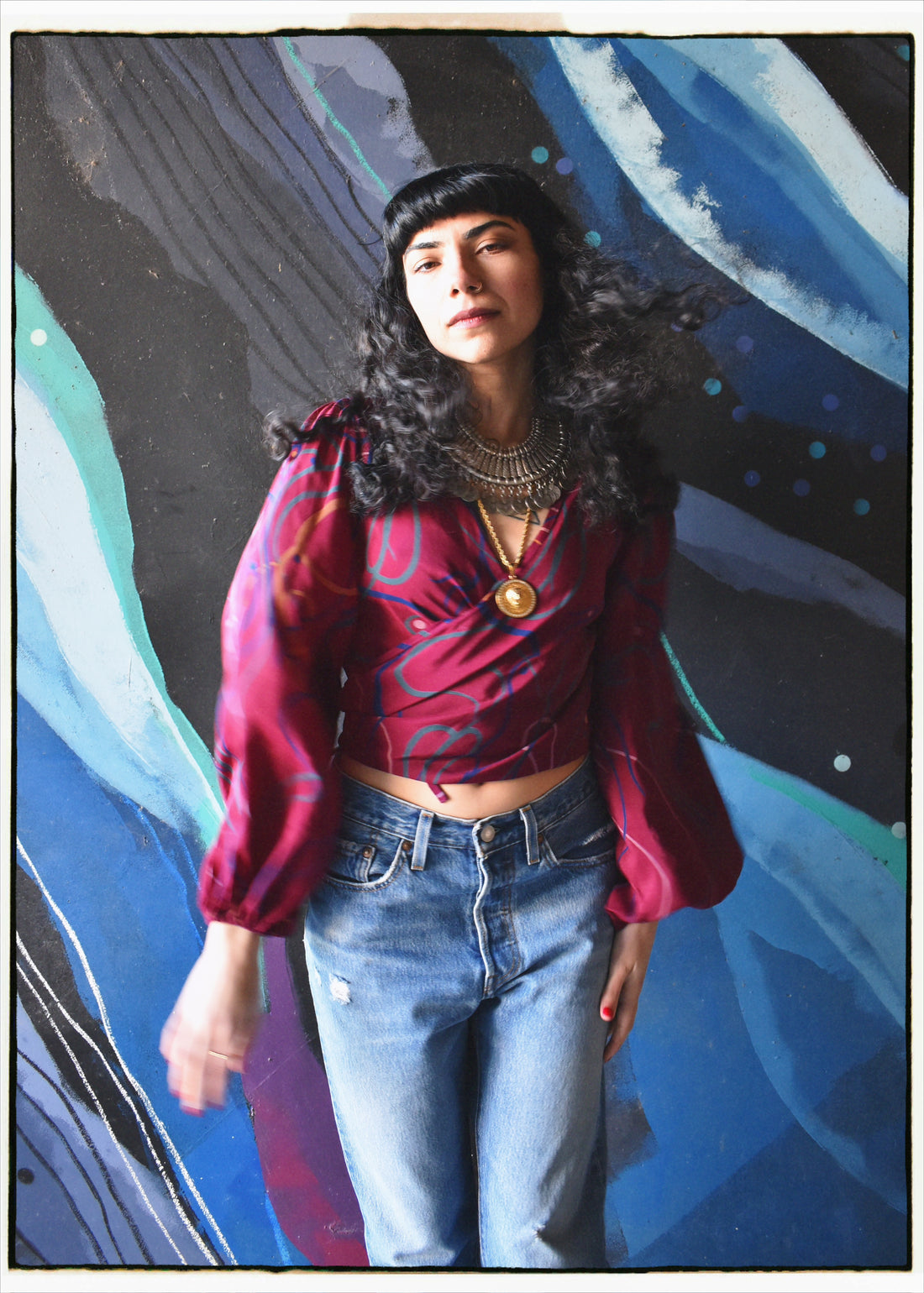 A woman with dark curly hair and a black beret stands against an abstract blue, black, and white painted background. She is wearing a shiny magenta wrap-style long-sleeve crop top with blue abstract lines, layered gold necklaces, and light-wash blue jeans.