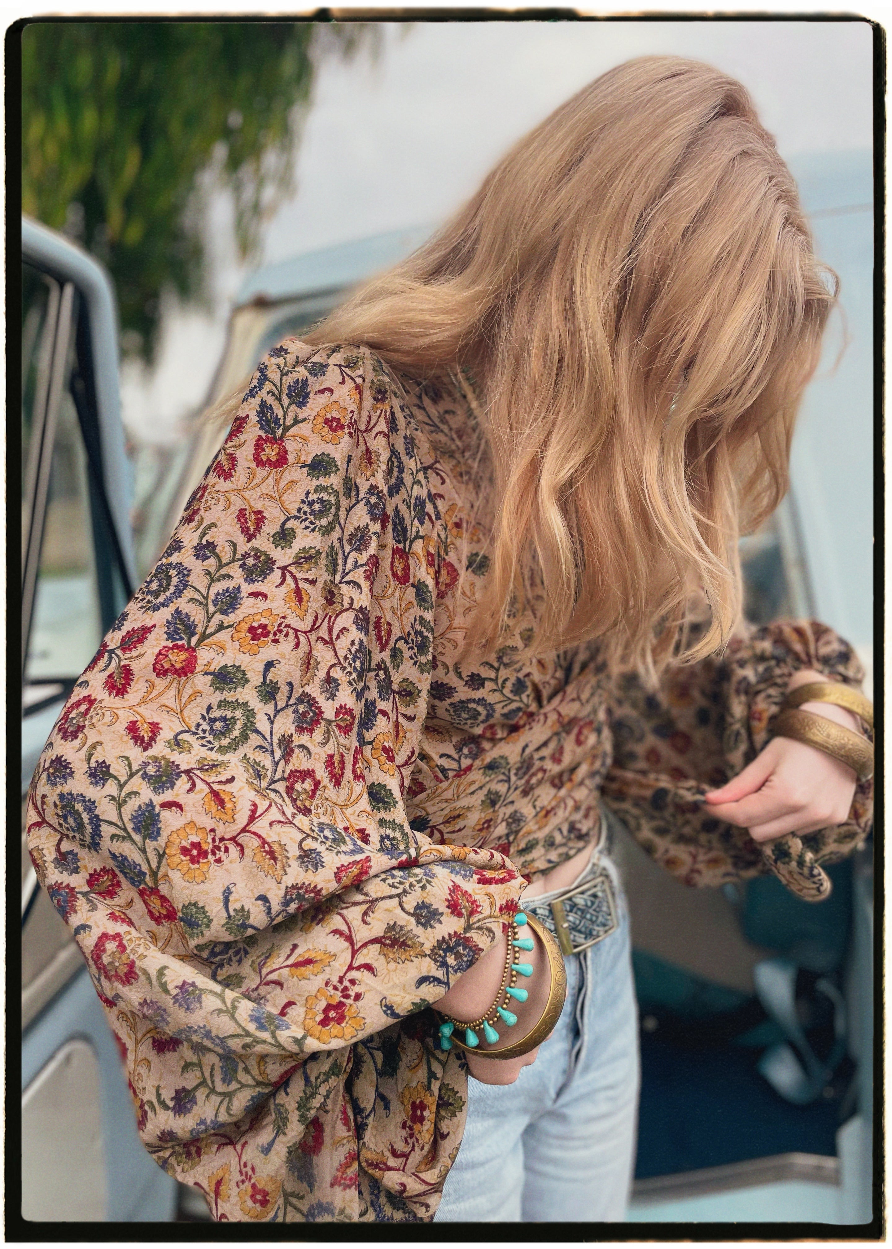 Woman wearing a bohemian style balloon sleeved blouse made from vintage Indian repurposed silk crepe saree sitting in the back of a vintage car with a scenic background