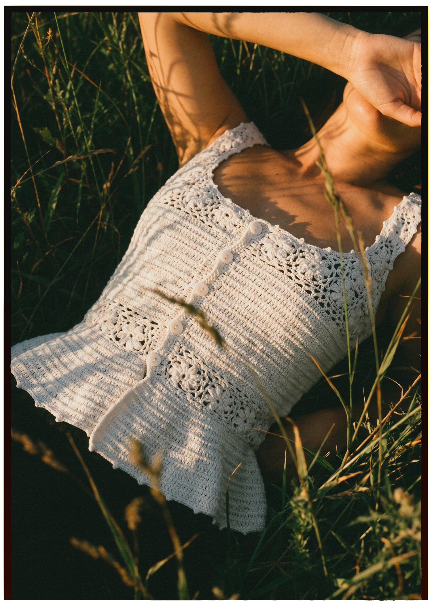 A woman lying in tall grass outdoors, wearing a white crocheted sleeveless top with lace trim and ruffled edges. Warm sunlight casts shadows across her body and the garment.