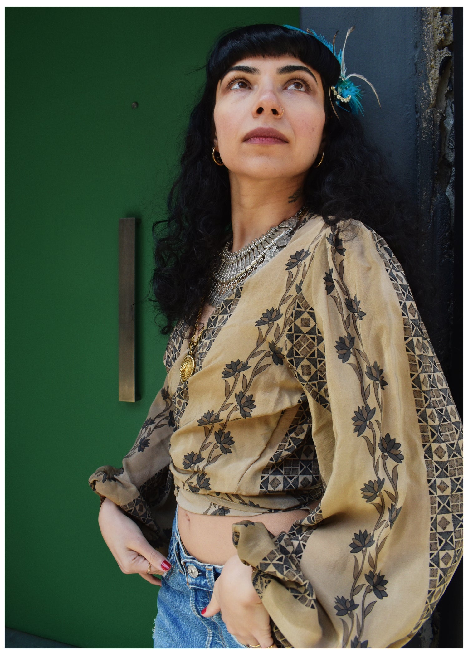 A woman with dark curly hair and a feather headpiece wears a beige bohemian-style blouse with black floral and geometric patterns, paired with blue jeans, posing against a dark green door.