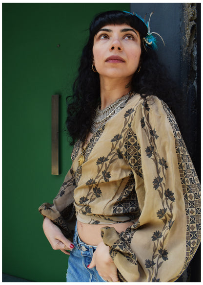 A woman with dark curly hair and a feather headpiece wears a beige bohemian-style blouse with black floral and geometric patterns, paired with blue jeans, posing against a dark green door.