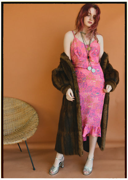 A woman stands in a studio wearing a bright pink patterned midi dress with spaghetti straps, a dark brown faux fur coat draped over her shoulders, layered gold necklaces, and metallic silver high-heeled sandals. A woven round chair is visible on the left against a solid warm orange-brown backdrop.