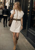 Woman in a white dress with a belt and brown boots walking on a street.