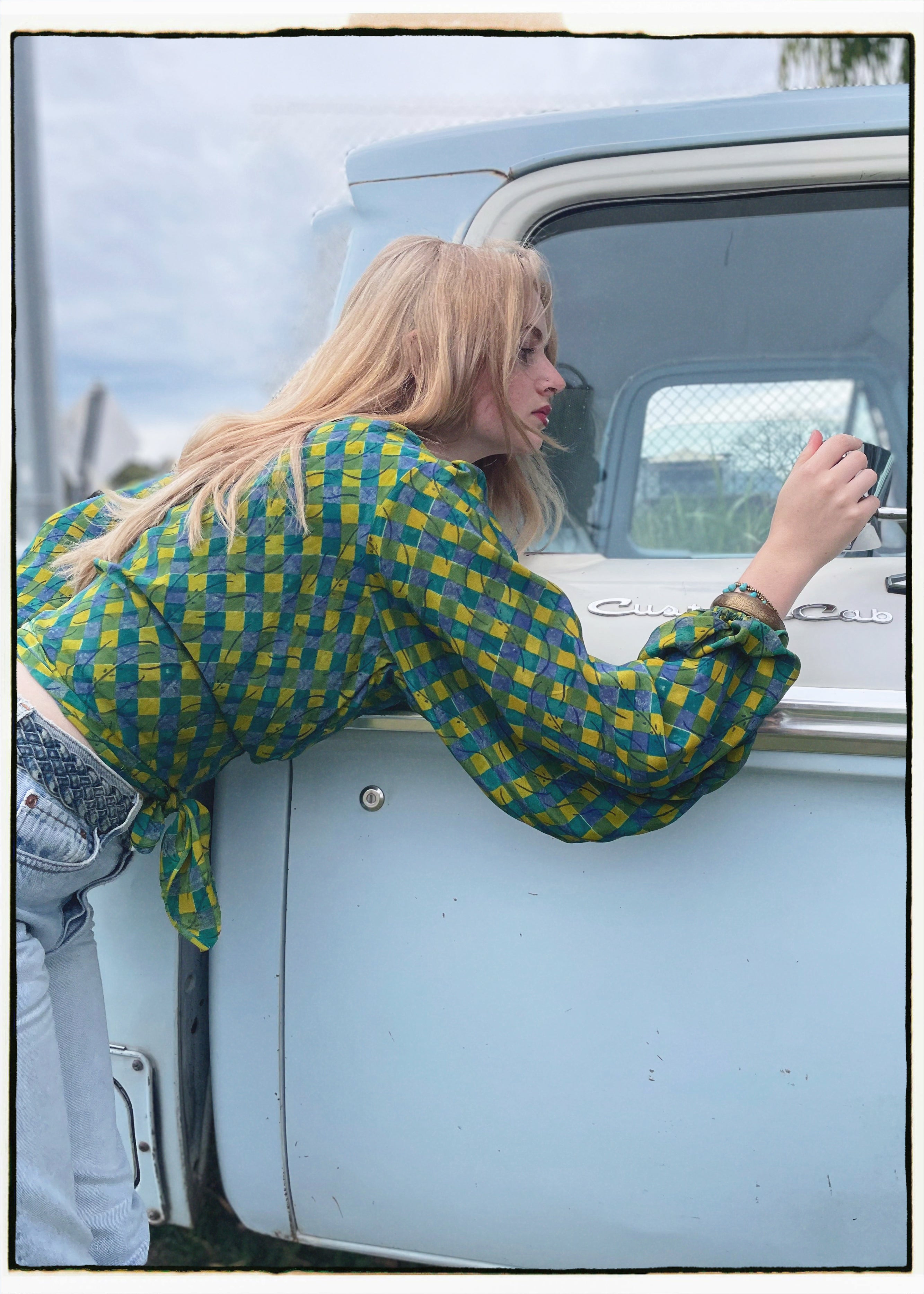 Woman wearing a bohemian style balloon sleeved blouse made from vintage Indian repurposed silk crepe saree sitting in the back of a vintage car with a scenic background
