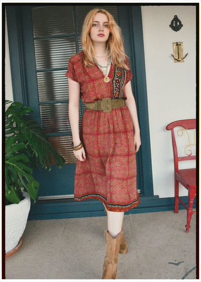 Woman wearing a red patterned dress with a belt, standing in a room with a plant and chair.