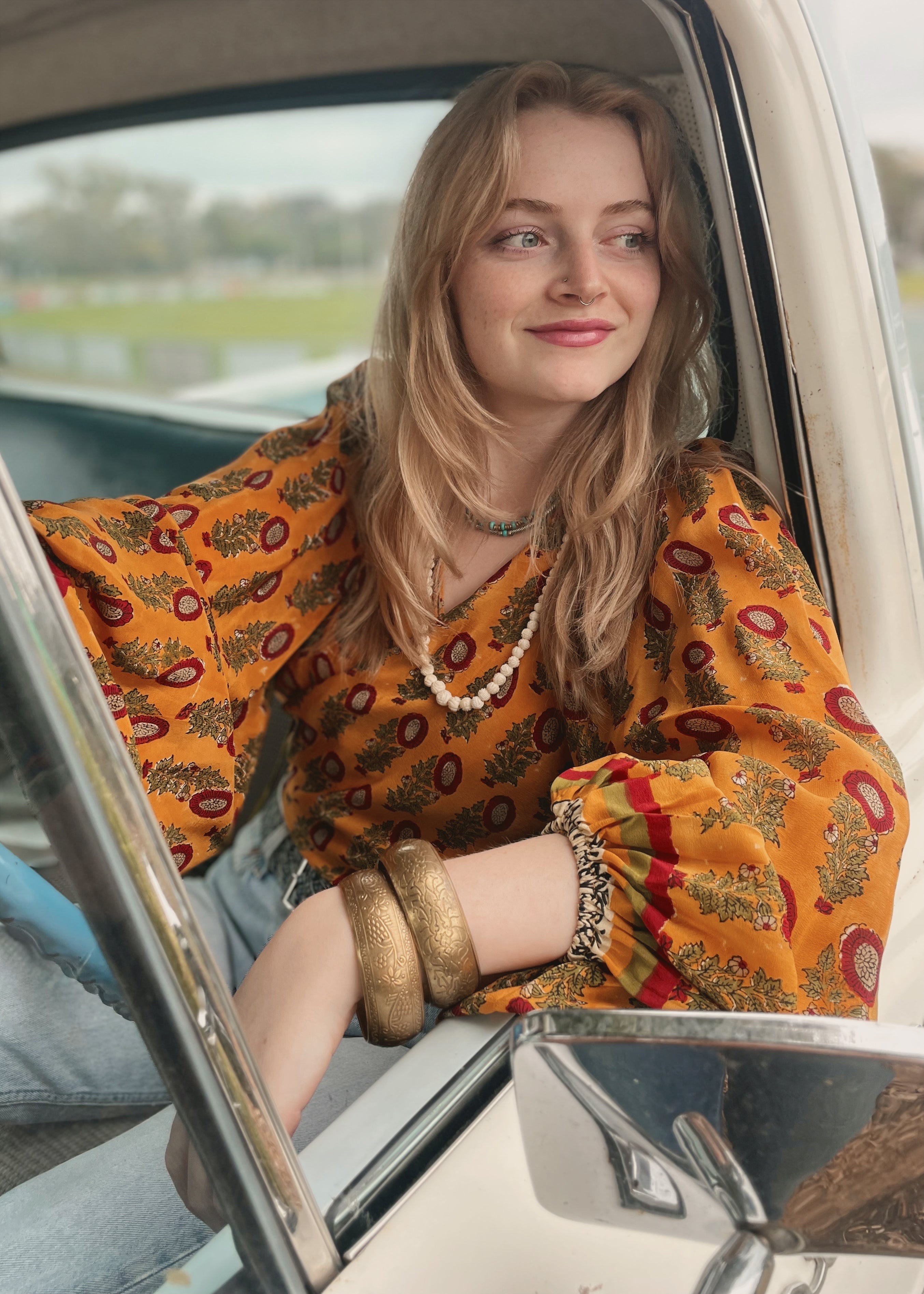 Woman wearing a bohemian style balloon sleeved blouse made from vintage Indian repurposed silk crepe saree sitting in the back of a vintage car with a scenic background