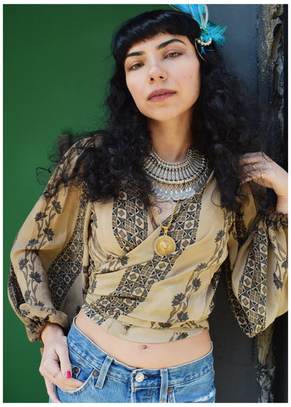 A woman with long black curly hair and a blue feather accessory leans against a dark textured wall, wearing a beige bohemian patterned blouse with blue jeans and layered silver necklaces.