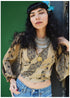 A woman with long black curly hair and a blue feather accessory leans against a dark textured wall, wearing a beige bohemian patterned blouse with blue jeans and layered silver necklaces.